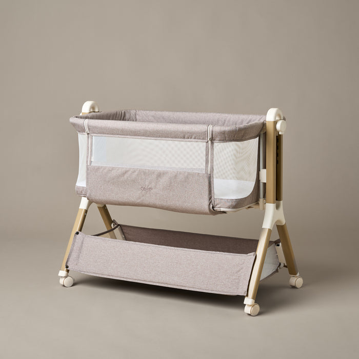 Bedside crib with taupe woven fabric, mesh sides, zipped up parent window, wood effect frame with ivory plastics & wheels. Storage basket underneath.