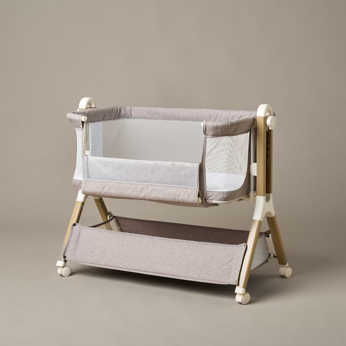 Bedside crib with taupe woven fabric, mesh sides, unzipped  parent window, allowing easy access to baby, wood effect frame with ivory plastics & wheels. Storage basket underneath.