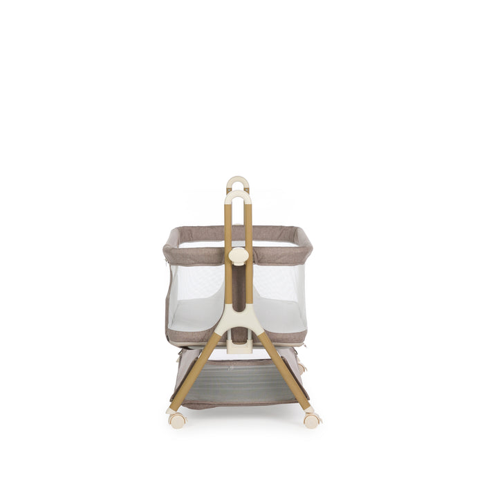 Side view of Bedside crib with taupe woven fabric, mesh sides, zipped up parent window, wood effect frame with ivory plastics & wheels. Storage basket underneath. Crib on its lowest setting.