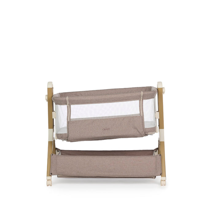Bedside crib with taupe woven fabric, mesh sides, zipped up parent window, wood effect frame with ivory plastics & wheels. Storage basket underneath. Cirb on a tilt, a feature to help with colic