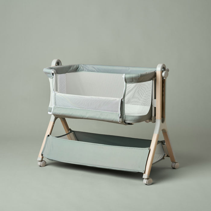 Bedside crib with sage fabrics, on a wood effect frame, showing side panel unzipped allowing parent easy access to newborn.