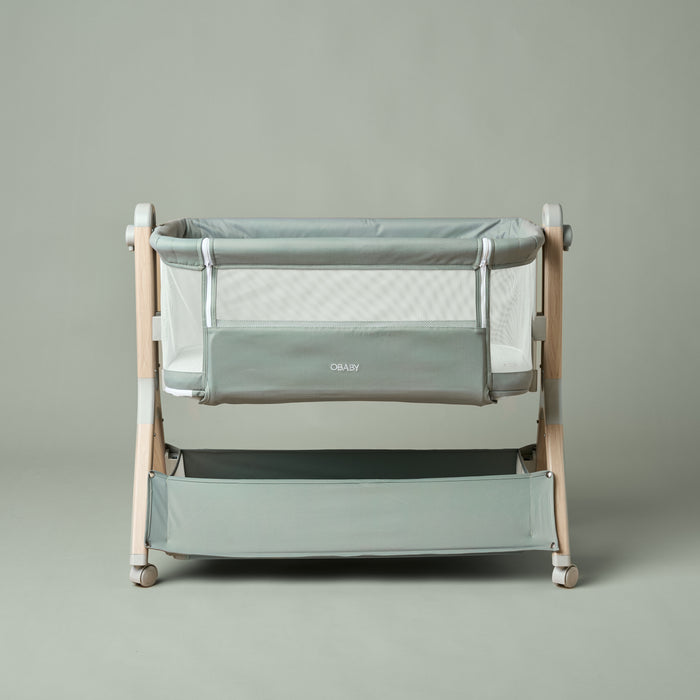 Bedside Crib with sage green fabric, wood effect metal frame, with grey wheels & grey casters, zipped up front parent panel & storage basket at the bottom.
