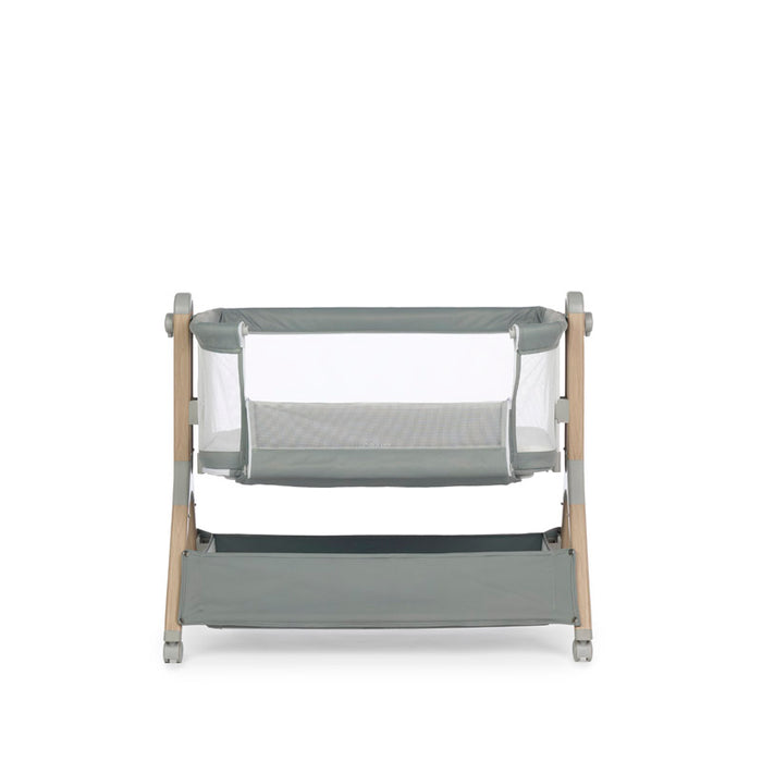 Bedside crib with sage green fabric, mesh sides, wood effect frame & grey plastics & casters, image shows parent window open, allowing easy parent access to the newborn. Storage basket underneath crib