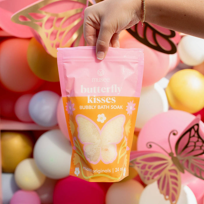 Hand holding a pink and orange package of 'butterfly kisses' bubble bath soak with colorful balloons in the background.