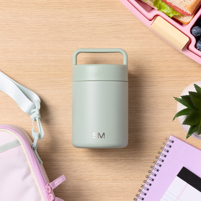 Flat lay of sage green insulated food container with SM logo surrounded by pink notebook, lunch box with sandwich and blueberries, plant leaves, and pale pink pouch on light wood surface.