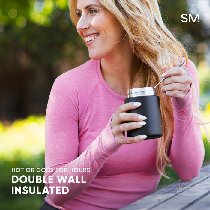 Woman holding black insulated beverage container with metal straw in outdoor garden setting, wearing pink long-sleeved shirt. Simple Modern double-wall insulated drinkware product photography.