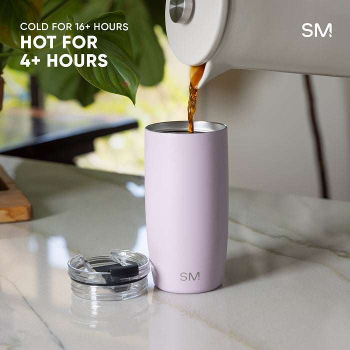 Pale lavender-pink insulated tumbler with stainless steel lid being filled with amber beverage from white carafe. Text reads "Cold 16+ Hours" and "Hot 4+ Hours." Modern kitchen setting with marble countertop and green