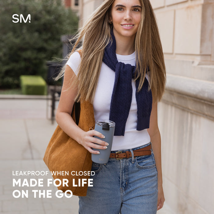 Woman holding gray Simple Modern Voyager tumbler wearing navy scarf and tan tote bag in outdoor urban setting. Text reads: "Leakproof When Closed, Made for Life, On the Go."